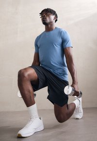 Αθλητικός άντρας που φοράει ανοιχτό μπλε κοντομάνικο πουκάμισο και μαύρες βερμούδες, εκτελεί μια προβολή κρατώντας αλτήρες και στις δύο παλάμες, σε ομαλή επιφάνεια.