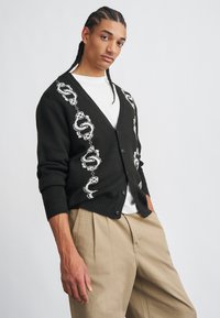 Jeune homme avec des tresses en cornrows portant un cardigan noir à boutons avec un motif de chaînes et de dés blancs, une chemise blanche et un pantalon beige, posant de côté.