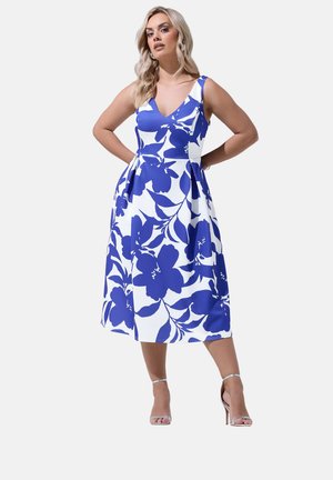 Frau in einem ärmellosen weißen Midikleid mit großem blauem Blumenprint, die mit den Händen hinter dem Rücken steht, silberne Sandalen mit hohen Absätzen.