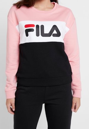 Femme portant un sweat-shirt FILA rose, blanc et noir et un pantalon noir, debout avec les bras détendus le long du corps.