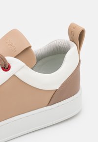 Baskets beige et blanches avec une tige en cuir texturé, un col blanc lisse, des œillets rouges et une semelle en caoutchouc blanche plate.