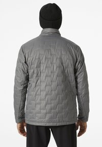 Grijze geïsoleerde jas met een gewatteerd patroon, hoge kraag en lange mouwen. Zwarte gebreide beanie gedragen, met een gladde textuur.