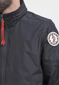 Mörkblå jacka för män, tillverkad av slät tyg, med hög krage, röd dragkedja och ett runt märke på ärmen med text.