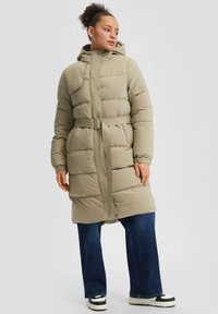 Puhasti plašč v svetlo zeleni barvi, srednje dolžine s kapuco, s pasom, z gumbi na zaklopu in kviltano teksturo. Kombiniran z širokimi kavbojkami in supergami.