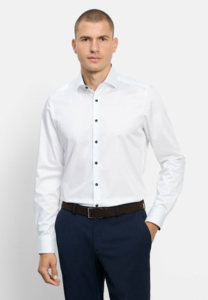 Mann trägt ein weißes Hemd mit langen Ärmeln und schwarzen Knöpfen, marineblaue Anzughose und einen dunkelbraunen Gürtel vor einem neutralen Hintergrund.