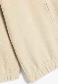 Veste en polaire beige avec une texture douce, dotée de poignets élastiques et d'une fermeture éclair sur toute la longueur. Montre un gros plan de la surface du tissu.