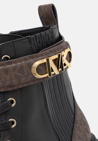 Bottine en cuir noir avec tissu marron à motifs, ferrures logo dorées et accents côtelés sur la tige. Modèle à lacets inclus.