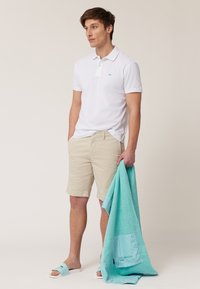 Witte polo, beige short, en blauwe slippers. Houdt een turquoise handdoek met een gestreepte acccent vast. Eenvoudige, casual zomeroutfit.