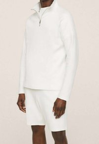 Pullover bianco con zip e pantaloni corti abbinati, realizzati in tessuto morbido, con maniche lunghe e vestibilità rilassata. Nessun motivo o accento visibile.