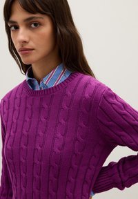 Lila, kabelstrickpullover mit Rundhalsauschnitt, strukturiertem Muster und gerippten Bündchen, über einem blau-weißen gestreiften Hemd mit Kragen getragen.