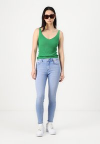 Groen geribbeld tanktop, lichtblauwe skinny jeans en witte sneakers. De outfit heeft een aansluitend ontwerp en een gladde textuur. Bruine oversized zonnebril.
