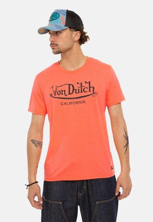 Homme portant un t-shirt orange Von Dutch California, une casquette trucker bleue et noire, un jean foncé, montrant des tatouages sur les bras, regardant à gauche.