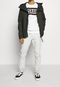 Svart jacka med dragkedja och huva över en vit sweatshirt med vinröd text. Ljusgrå byxor och svarta sneakers med vita sulor fulländar outfiten.