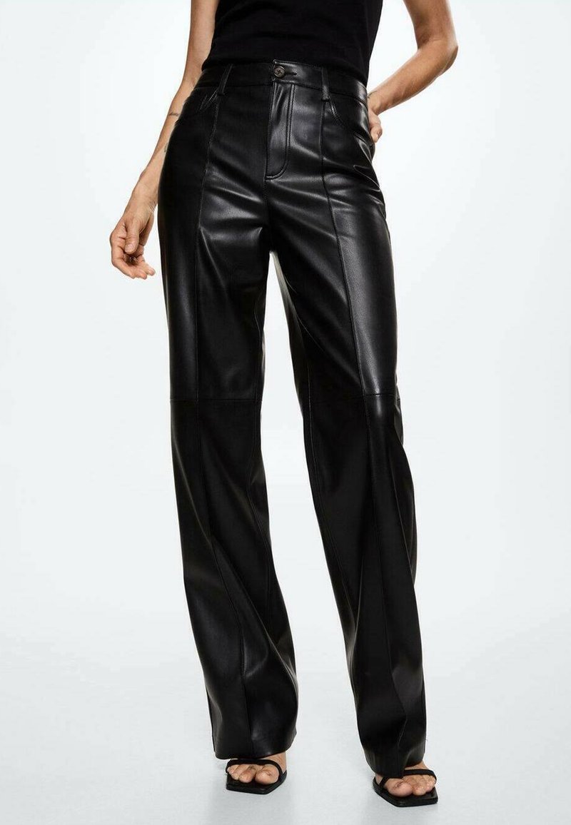 Femme portant un pantalon en cuir noir taille haute à jambes droites avec coutures avant et des talons ouverts noirs, debout devant un fond blanc.