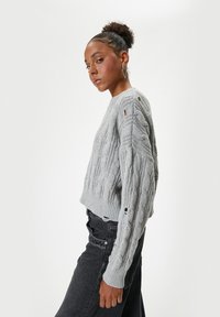 Grijze kabelgebreide trui met een cropped ontwerp, voorzien van textuurpatronen en kleine uitsparingen op de mouwen. Gedeeld met donkere jeans.