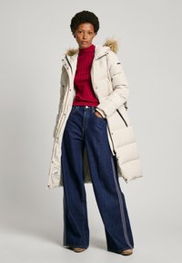 Beige vadderad jacka med en huva klädd i päls, röd ribbad polotröja och vida blå jeans med dekorativ sömnad på sidorna.