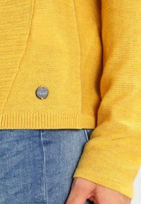 Gelber Strickpullover mit strukturierter Oberfläche, abgerundetem Saum und einem metallischen Logo-Detail in der Nähe der Naht. Über Denim-Stoff getragen.