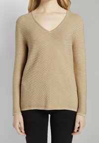 Beige V-Neck-Pullover mit ribbeliger Textur, mit engem Schnitt und langen Ärmeln, kombiniert mit schwarzen Hosen.