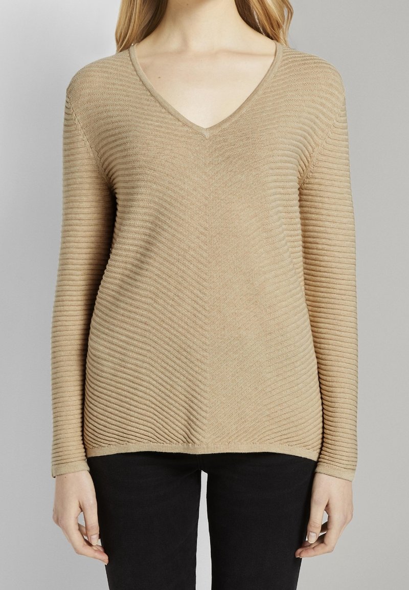 Beige V-Neck-Pullover mit ribbeliger Textur, mit engem Schnitt und langen Ärmeln, kombiniert mit schwarzen Hosen.