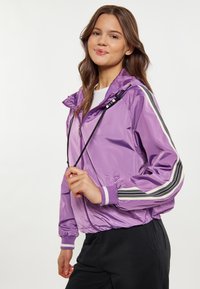 Giacca leggera viola con cappuccio, caratterizzata da strisce bianche e nere sulle maniche, polsini elastici e coulisse regolabile.