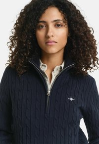 Marineblauer Pullover mit Kabelstrickmuster, halbzipper Kragen und silbernem Reißverschluss. Mit kleinem Logo auf der Brust und einer weichen Textur.