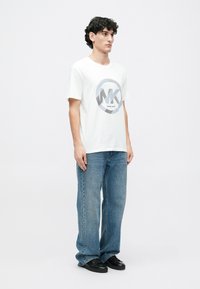 Camiseta blanca de manga corta con un logo gráfico circular en gris y azul. Combinada con unos vaqueros sueltos de corte ligero en azul claro y zapatillas negras.