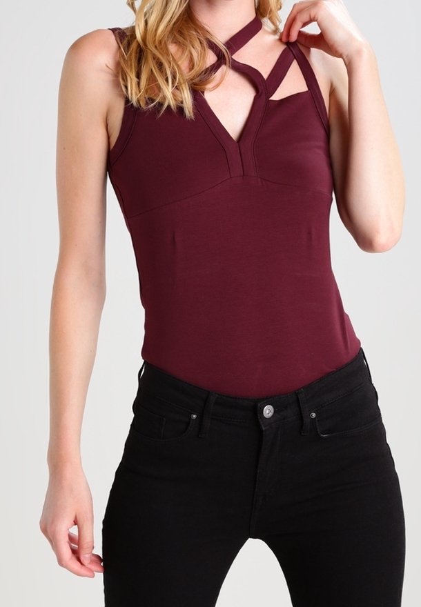 Femme portant un haut bordeaux foncé sans manches avec un design de bretelle asymétrique et un jean noir taille haute, main touchant la bretelle de l'épaule.