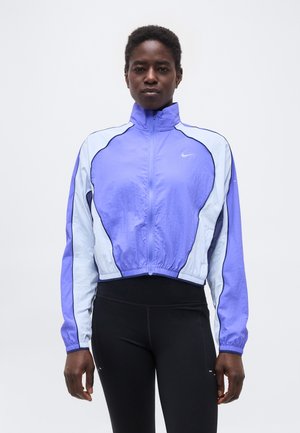 Άτομο που στέκεται, φορώντας αθλητικό μπουφάν με φερμουάρ σε μωβ και άσπρο και μαύρα κολάν, σε απλό λευκό φόντο.