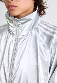 Zilveren Adidas-jack met een hoge kraag, voorzien van een rits aan de voorkant en drie witte strepen op de schouders. Glad, reflecterend materiaal.