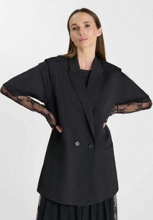 Frau mit langen braunen Haaren, die einen übergroßen schwarzen Blazer mit transparenten Spitzenärmeln und einen passenden schwarzen Spitzenrock vor einem schlichten Hintergrund trägt.