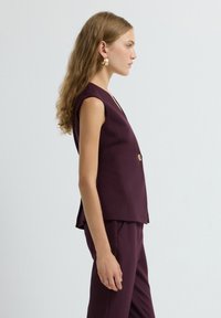 Gilet senza maniche bordeaux con scollo a V profondo, caratterizzato da una chiusura con un singolo bottone dorato, completato da un tessuto liscio e structuro.