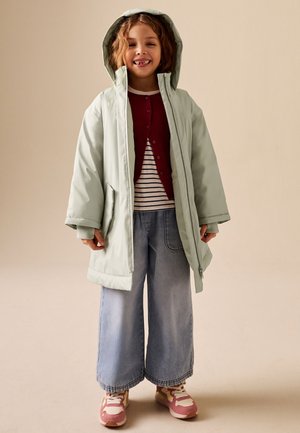 Unge barn iført en lysegrøn hættetrøje, rød cardigan med knapper, stribet skjorte, jeans med vide ben og lyserøde sneakers, der smiler.