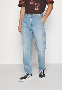 Pantalones cargo de mezclilla azul claro con un ajuste relajado, costuras visibles, bolsillos laterales y una textura ligeramente desgastada. Combinados con zapatillas negras y blancas.