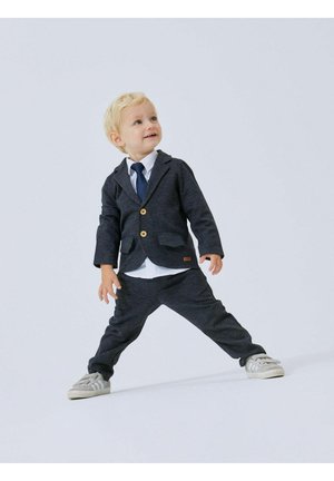 Blazer grigio con bottoni in legno, camicia bianca, cravatta blu e pantaloni grigi. Tessuto leggero, vestibilità rilassata e scarpe in stile sneaker.
