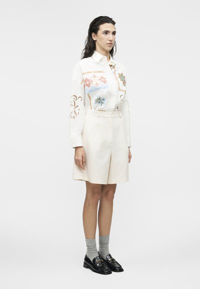 Femme debout avec des cheveux effet mouillé, portant un chemisier blanc à manches longues avec des motifs colorés, un short crème, des chaussettes grises et des mocassins noirs.