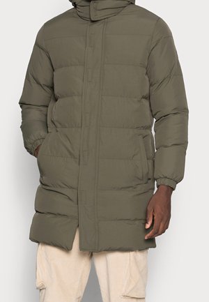 Hombre con un abrigo acolchado verde oliva con bolsillos laterales y puños elásticos, combinado con pantalones de pana beige.