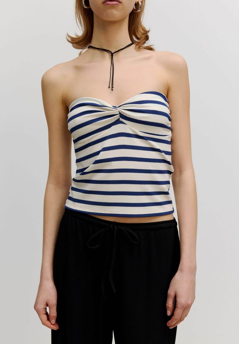 Strapless ribbeltop met marineblauwe en witte horizontale strepen, voorzien van een twistdetail in het midden. Draagt met zwarte broek met trekkoord.