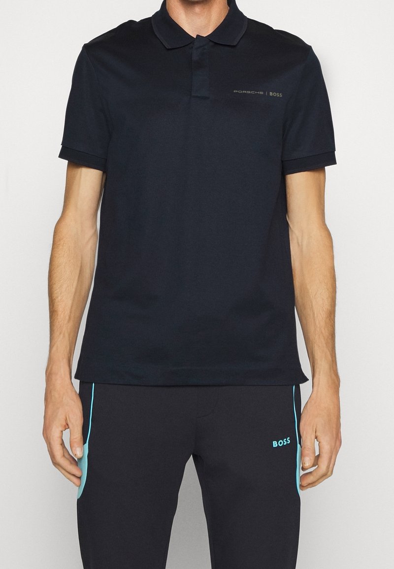 Polo shirt bleu marine en tissu texturé, col à deux boutons, et logo discret sur la poitrine. Assorti avec un pantalon foncé comportant des accents aqua.
