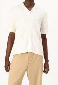 Wit shirt met korte mouwen, voorzien van een gestructureerd golfpatroon, knoopsluiting en een kraag. Gecombineerd met lichtbeige broek.