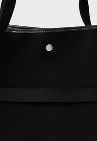 Μαύρη τσάντα tote από μαλλί με μαύρη πλεκτή λαβή, μαύρες δερμάτινες λεπτομέρειες με λευκή ραφή και ένα μικρό στρογγυλό χρυσό κουμπί στο μπροστινό μέρος.