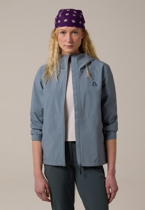 Mujer con cabello largo y rubio, que lleva un pañuelo morado con flores en la cabeza, chaqueta azul claro, camisa blanca y pantalones oscuros, de pie frente a un fondo neutro.