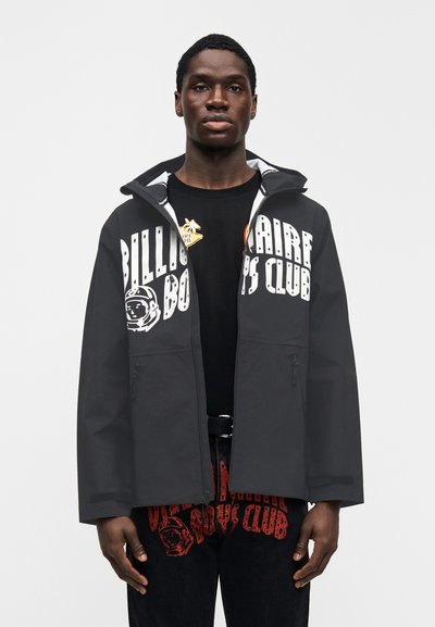 Σκούρα γκρι αδιάβροχο μπουφάν με κουκούλα, διακοσμημένο με λευκό κείμενο και γραφικά στοιχεία. Τσέπες με φερμουάρ και άνετη εφαρμογή. Φορεμένο πάνω από μαύρο πουκάμισο.