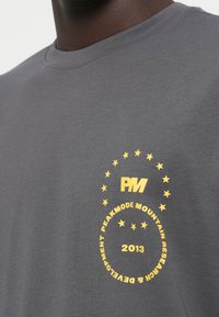 Hallide hall t-särk, millel on kollane graafiline kujundus, kus on kirjas "PM", tähed ja "PEAKMODE MOUNTAIN RESEARCH & DEVELOPMENT 2013". Lühikesed varrukad.