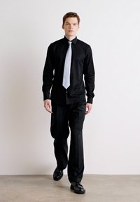Juodas marškiniai ir kelnės, derinamos su šviesiai pilku kaklaraiščiu. Batai juodi. Audinys atrodo lygus ir pritaikytas, suteikdamas elegantišką išvaizdą.