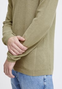 Pulover texturat de culoare verde măslinie, cu manșete și tiv ribate, având un model orizontal subtil. Puratat peste blugi albaștri deschiși, cu mâinile poziționate pe mâneci.