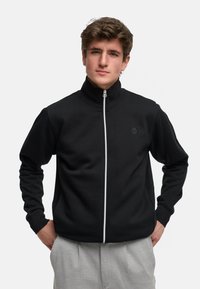 Svart zip-up jakke med høy krage og hvit glidelås, laget av mykt stoff, med lange ermer og en subtil logo på brystet.