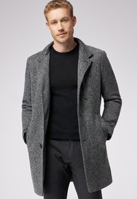 Grau gestreifter Mantel mit Reverskragen, seitlichen Taschen und einfacher Knopfverschluss. Kombiniert mit einem schwarzen Rundhalsoberteil und grauen Hosen.