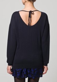 Marineblauer Pullover mit lockerem Schnitt, tiefem Rückenausschnitt und einer gebundenen Detail. Der Stoff wirkt weich, kombiniert mit einem strukturierten blauen Spitzenrock.