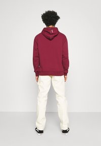 Person mit lockigem Haar, der einen bordeauxfarbenen Hoodie, off-white Hosen und schwarze Sneaker trägt und einer schlichten weißen Wand gegenübersteht.