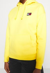 Felpa gialla con tasca frontale, dotata di lacci e un piccolo logo "TOMMY JEANS" multicolore sul petto, realizzata in tessuto morbido.